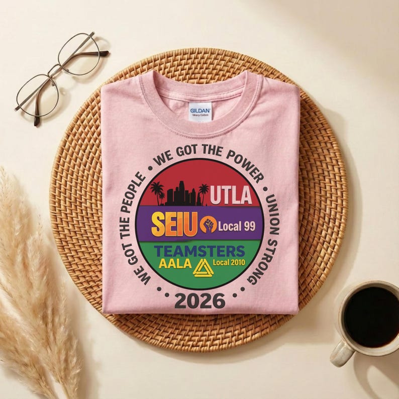 May include: A pale pink t-shirt with a circular graphic. The graphic includes the text "WE GOT THE POWER", "UNION STRONG", "UTLA", "SEIU Local 99", "TEAMSTERS AALA Local 2010", and the year "2026". The t-shirt is on a woven mat.