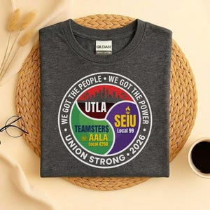 May include: A dark gray, crew neck t-shirt with a circular graphic featuring the text "WE GOT THE PEOPLE, WE GOT THE POWER, UNION STRONG 2026." The graphic includes the logos for UTLA, Teamsters AALA Local 4200, and SEIU Local 99.