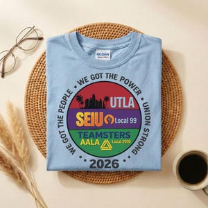 May include: Pale blue Gildan Heavy Cotton t-shirt with a circular graphic. The graphic includes the text "WE GOT THE POWER", "UTLA", "SEIU Local 99", "TEAMSTERS AALA Local 2010", and "2026". The shirt is on a woven mat.