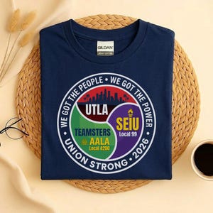 May include: Navy blue t-shirt with a round graphic featuring the text "WE GOT THE PEOPLE WE GOT THE POWER" and "UNION STRONG 2026". The graphic includes the logos of UTLA, Teamsters, AALA, and SEIU.
