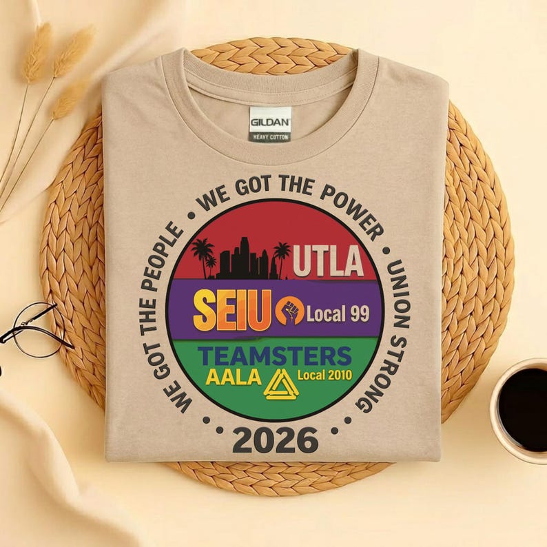 May include: A beige t-shirt with a circular design. The design includes the text "WE GOT THE POWER" and "UNION STRONG." The graphic features "UTLA," "SEIU Local 99," and "TEAMSTERS AALA Local 2010." The year "2026" is also printed on the shirt.