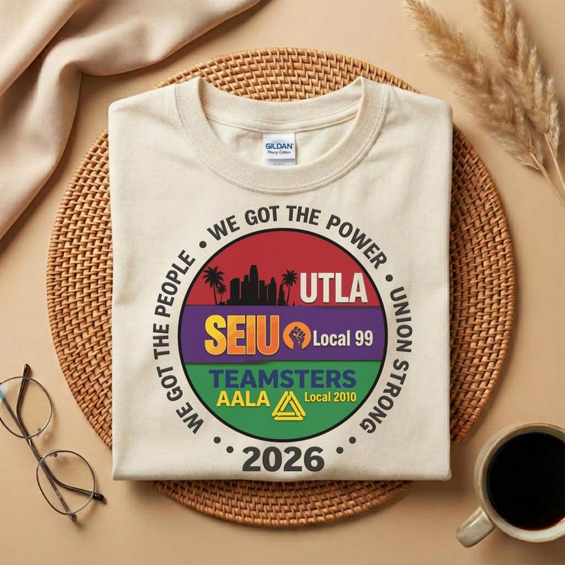 May include: A cream-coloured t-shirt with a circular graphic. The graphic includes the text "WE GOT THE POWER," "WE GOT THE PEOPLE," "UTLA," "SEIU Local 99," "TEAMSTERS AALA Local 2010," and "2026." A coffee cup and eyeglasses are visible.