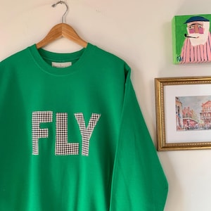 May include: A bright green sweatshirt with the word "FLY" appliqued in a black and white checkered pattern. The sweatshirt is hanging on a wooden hanger against a white wall. A small painting and framed print are visible in the background.
