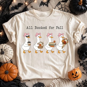Puede incluir: Una camiseta color crema con el texto All Booked for Fall y un gráfico de cuatro patos blancos con lazos rosas. Los patos llevan una taza de café, una cesta de calabazas, una pila de libros y un ramo de flores de otoño.