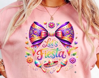 May include: Pink t-shirt with a colorful "Let's Fiesta" graphic. The design features a large purple bow, flowers, maracas, and a sombrero. The text is in rainbow colors, creating a festive and vibrant look.