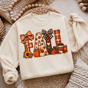 May include: A cream-colored crewneck sweatshirt featuring the word FALL in large, decorative letters. The letters are filled with patterns of plaid, autumn leaves, pumpkins, sunflowers, hearts, and a coffee cup, accented with bows and buttons.
