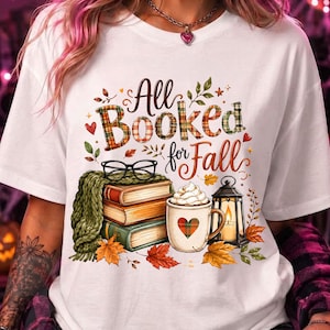 Op de afbeelding: Een wit t-shirt met een grafische print met de tekst All Booked for Fall. Het ontwerp bevat een stapel van drie boeken, een zwarte bril, een groene gebreide sjaal, een mok met een hartje, een lantaarn en kleurrijke herfstbladeren.