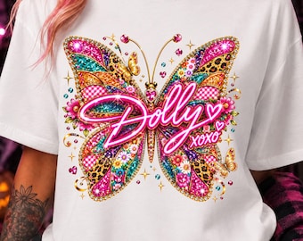 May include: A white t-shirt featuring a large, colorful butterfly graphic with neon pink text that reads Dolly XOXO. The butterfly wings are decorated with various patterns including leopard print, floral motifs, and glittery pink, gold, and teal accents.