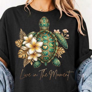 May include: A black t-shirt featuring a graphic of a green sea turtle surrounded by white and yellow plumeria flowers and gold fern leaves. The text "Live in The Moment" is printed in a gold cursive script below the turtle illustration.