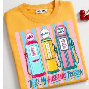 May include: Mustard yellow sweatshirt with a graphic of three vintage gas pumps in blue, yellow, and pink, set against a striped background. The text "That's My Husband's Problem" is at the bottom.