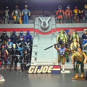 May include: A display of G.I. Joe action figures on shelves. The figures are diverse in colour and size, representing soldiers and other characters. The G.I. Joe logo is in the centre. The figures are arranged in rows.