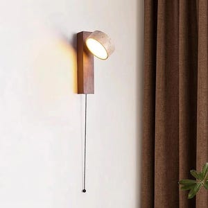 May include: A modern wall-mounted lamp with a rectangular wooden base and a cylindrical lampshade. The lamp is turned on, emitting a warm light. A black chain hangs down, likely for turning the lamp on and off. The lamp is mounted on a white wall.