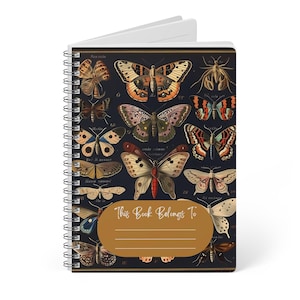 May include: A spiral-bound notebook with a dark blue cover displaying vintage butterfly illustrations in brown, orange, and cream. The notebook features the text "This Book Belongs To" on a gold label, ideal for notes or journaling.
