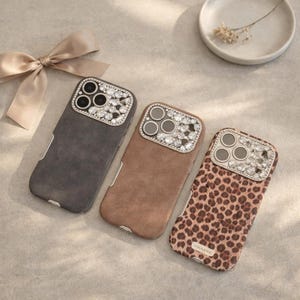 Puede incluir: Tres fundas para teléfono con cubiertas de cámara enjoyadas. Las fundas son grises, marrones y con estampado de leopardo. Las fundas tienen una textura similar a la gamuza. Un lazo beige y un plato blanco pequeño con flores secas están en el fondo.