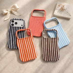 Puede incluir: Una colección de fundas para teléfonos con patrones de rayas verticales. Las fundas son de varios colores, incluyendo azul marino, naranja, coral, marrón y azul claro. También hay un lazo blanco y un pequeño cuadrado decorativo con flores secas.