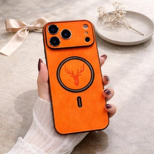 Könnte beinhalten: Orangefarbene Handyhülle mit einem Hirschkopf-Emblem. Die Hülle hat einen schwarzen Rand und ein kreisförmiges Design in der Mitte. Sie hat Aussparungen für die Kamera des Telefons und den Ladeanschluss.