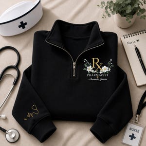 Könnte beinhalten: Ein schwarzes Sweatshirt mit kurzem Reißverschluss, verziert mit einem goldenen Rx-Symbol, umgeben von weißen Blüten und grünen Blättern, sowie dem Schriftzug PHARMACIST und Amanda Garcia. Der Ärmel zeigt ein goldenes Stethoskop und eine Herzlinie.
