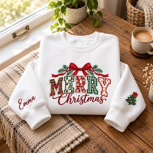 Peut inclure: Un sweat-shirt blanc à col rond orné d'un motif central avec un nœud rouge, des branches de pin vertes et le texte Merry Christmas dans des motifs à carreaux, flocons et pois. Le prénom Emma est imprimé sur une manche et un sapin sur l'autre.
