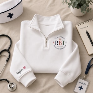 May include: A white quarter-zip sweatshirt featuring a circular logo on the chest that reads Registered Behavior Technician with the letters RBT in the center. The left sleeve is personalized with the name Taylor and a small pink heart icon.