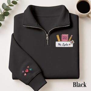 Könnte beinhalten: Ein schwarzes Sweatshirt mit kurzem Reißverschluss und einer Bruststickerei, die ein Lineal, ein rosa Notizbuch mit der Aufschrift MATH NOTES, einen Bleistift und ein weißes Etikett mit der Aufschrift Ms. Lydia sowie eine Blume und ein Herz zeigt. Kleine bunte Herzen verzieren den Ärmel.