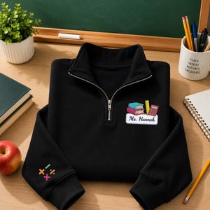 Puede incluir: Una sudadera negra con cremallera corta y un parche bordado en el pecho con dos libros, una regla, una calculadora y el texto Ms. Hannah. La manga izquierda muestra símbolos matemáticos bordados en colores, incluyendo signos de suma y división.