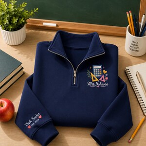 Könnte beinhalten: Ein marineblaues Sweatshirt mit kurzem Reißverschluss und einer Stickerei auf der Brust, die einen Taschenrechner, Lineale, Blumen und den Text "Mrs. Johnson" zeigt. Der Ärmel ist mit dem Text "Math Teacher EST. 2023" und zwei kleinen roten Herzen bestickt.