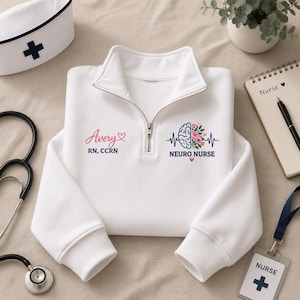 Op de afbeelding: Een wit sweatshirt met een kwartritssluiting en een afbeelding van een brein met bloemendetails en een ECG-lijn. De tekst luidt Avery, RN, CCRN en NEURO NURSE. Het ontwerp bevat roze hartjes en blauwe letters op de voorzijde van het kledingstuk.