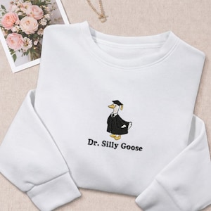 May include: White sweatshirt with a crew neck featuring an embroidered design of a goose wearing a graduation cap and gown. The text "Dr. Silly Goose" is below the goose. A bouquet of pink flowers and a necklace are in the background.