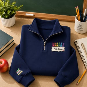 May include: A navy blue quarter-zip sweatshirt featuring a graphic of six colourful crayons above a piece of lined paper with the text "Mrs. Taylor" and a small pink heart. The left sleeve displays a small graphic of a stack of books with a green worm.