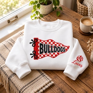 Könnte beinhalten: Ein weißes Sweatshirt mit Rundhalsausschnitt und einer Stickerei in Form eines rot-weiß karierten Wimpels mit dem Wort BULLDOGS in schwarzen Buchstaben. Auf dem rechten Ärmel befindet sich ein gesticktes rot-weißes Pompon-Motiv über dem Namen Lucas.