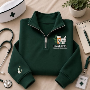 May include: A dark green quarter-zip sweatshirt featuring a graphic of a prescription bottle, mortar and pestle, and the text "David, CPhT PHARMACY TECHNICIAN". The sleeve includes a small embroidered capsule icon with a star above it.