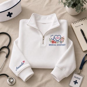 May include: A white quarter-zip sweatshirt featuring a graphic of a stethoscope shaped like a heart, a clipboard, and flowers, with the text "MEDICAL ASSISTANT" printed below. The sleeve is personalised with the name "Amanda" and a small pink heart.