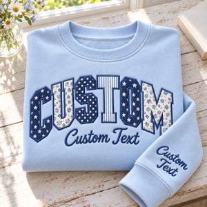 Könnte beinhalten: Ein hellblaues Sweatshirt mit Rundhalsausschnitt und dem Wort CUSTOM in großen, dunkelblauen Applikationsbuchstaben mit verschiedenen Mustern wie Sternen, Punkten, Streifen und Blumen. Der Text Custom Text ist darunter und auf dem Ärmel gestickt.