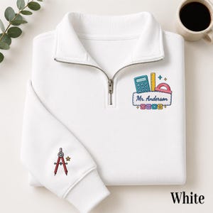 May include: A white quarter-zip sweatshirt featuring an embroidered design on the chest with a calculator, ruler, protractor, and the text "Mr. Anderson" above math symbols. The sleeve features an embroidered red drafting compass and a small star.
