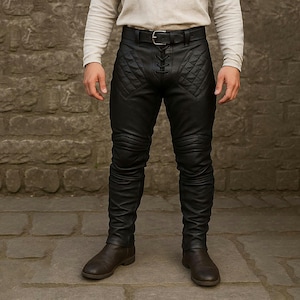 Könnte beinhalten: Schwarze Lederhose mit rautenförmiger Steppung an den Oberschenkeln und Knieschonern. Die Hose hat einen Schnürverschluss und einen Gürtel mit silberner Schnalle. Die Hose wird mit braunen Lederstiefeln kombiniert.