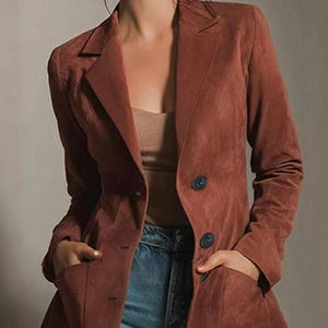 May include: A rust-colored blazer with a double-breasted design. The blazer has two rows of dark blue buttons and two front pockets. The model is wearing blue jeans.