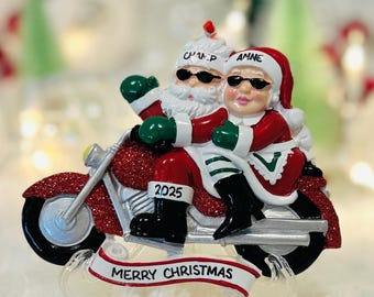 Puede incluir: Adorno navideño que representa a Papá Noel y Mamá Noel en una moto roja. Papá Noel lleva gafas de sol y un sombrero con la inscripción "Champ", mientras que Mamá Noel lleva gafas de sol y un sombrero con la inscripción "Amne". La moto muestra "2025" y una pancarta con "Merry Christmas".