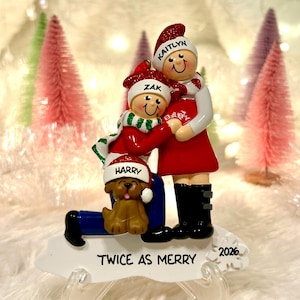 May include: A personalized Christmas ornament featuring three figures: a person named Kaitlyn, a person named Zak, and a brown dog named Harry. The figures wear red and white Santa hats and scarves. The base displays the text TWICE AS MERRY and 2026.