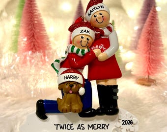 Könnte beinhalten: Ein Weihnachtsschmuck mit drei Figuren: eine Person namens Kaitlyn, eine Person namens Zak und ein Hund namens Harry. Sie tragen rot-weiße Mützen und Schals. Der weiße Sockel ist mit TWICE AS MERRY und der Jahreszahl 2026 beschriftet.