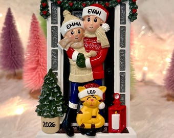 May include: A personalised Christmas ornament featuring two figures wearing hats labelled EMMA and EVAN, and a cat wearing a hat labelled LEO. The figures stand in a doorway with a green garland, a small tree marked 2026, and a red lantern.