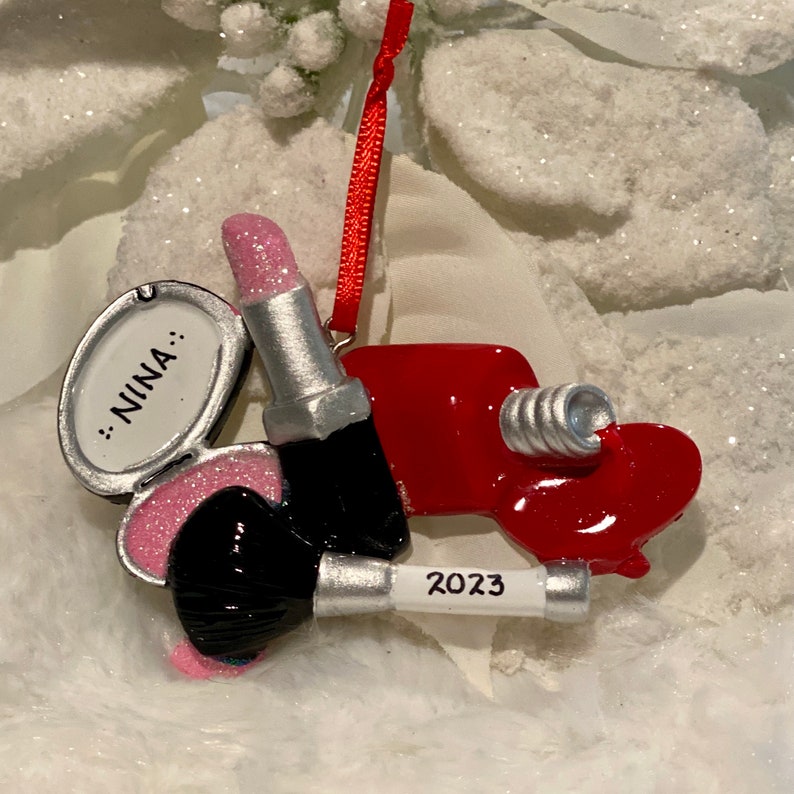 May include: A hanging ornament featuring a silver compact mirror with the name NINA, a glittery pink lipstick, a black makeup brush, and a red nail polish bottle with the year 2023 printed on the handle, all suspended by a thin red ribbon.