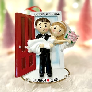 May include: A decorative ornament featuring a figurine of a person in a black tuxedo carrying another person in a white wedding dress through a red door frame. The text OCTOBER 10. 2026 is at the top, and LAUREN ❤️ CODY is printed at the base.