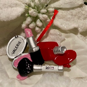 May include: A hanging ornament shaped like a makeup set featuring a red heart, a pink glittery lipstick, and a black makeup brush. The ornament includes a white oval mirror with the name VALERIE printed on it and a white handle labeled 2023.