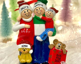 Könnte beinhalten: Ein weihnachtlicher Familienschmuck mit drei Figuren und einem Hund. Die Figuren tragen Mützen mit den Namen Jackie, Dominic und Luca. Der Hund trägt das Namensschild Kona. Ein rotes Geschenk trägt die Aufschrift Due Feb 2027. Auf der Hose steht 2026.