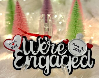 Könnte beinhalten: Ein schwarz-weißes Ornament mit dem Schriftzug We're Engaged in Schreibschrift. Ein weißes Herz auf der linken Seite zeigt die Namen Jack und Evie mit einem roten Herz. Ein weißer Kreis rechts zeigt das Datum 2. Juni 2026 mit kleinen roten Herzen.