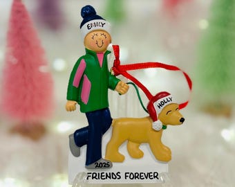 May include: A Christmas ornament featuring a figure wearing a dark blue hat labeled Emily, a green jacket, and dark blue pants, walking a tan dog wearing a red hat labeled Holly. The base is white with the text 2025 and FRIENDS FOREVER.