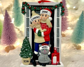 May include: A rectangular Christmas ornament featuring two figures wearing winter hats labeled Lindsey and Jake, and a cat labeled Honey. The figures stand in a doorway decorated with a green garland, a small Christmas tree marked 2026, and a red lantern.