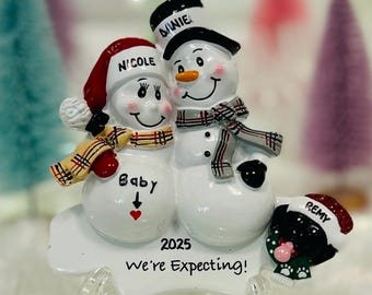 May include: A decorative ornament featuring two white snowmen and a black dog. The snowmen wear hats labeled Nicole and Daniel, with a plaid scarf on each. The dog wears a hat labeled Remy. Text reads 2025, Baby, and We're Expecting!