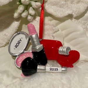 May include: A hanging ornament featuring a red lipstick tube, a black makeup brush with the year 2023 printed on the handle, and a silver compact mirror with the name RORY written in black text. The ornament hangs from a thin red ribbon.