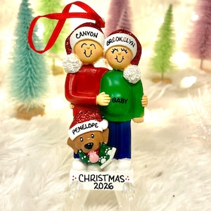 May include: A Christmas ornament featuring two figures and a dog wearing Santa hats. The hats are inscribed with the names Canyon, Brookelyn, and Penelope. The green shirt says BABY, and the base is inscribed with CHRISTMAS 2026.